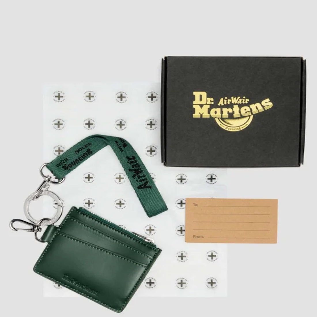 A Dr Martens Card Holder Keyring Green Kiev Leather Bag Charm with a matching strap and silver clasp rests next to a black Dr. Martens box, branded tissue paper, and a "To and From" brown gift tag on a light gray background.